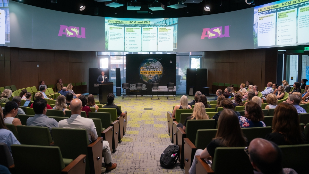 A group of people are seated in an auditorium facing a speaker and video screens. 